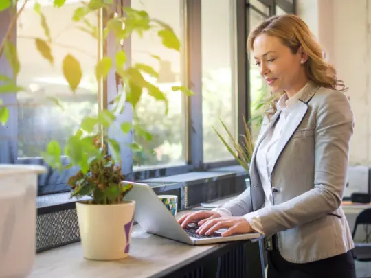 Mature businesswoman standing near windows and using laptop in modern office